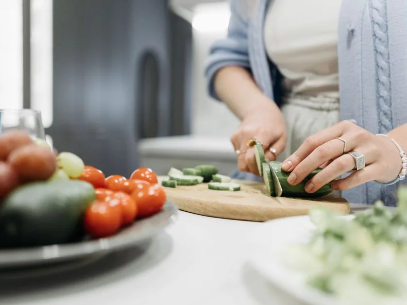 Mains qui tranchent un concombre sur une planche en bois, entourées de légumes frais